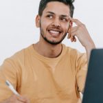 Work Future - Smiling young bearded Hispanic male entrepreneur thinking over new ideas for startup project and looking away dreamily while working at table with laptop and taking notes in notebook