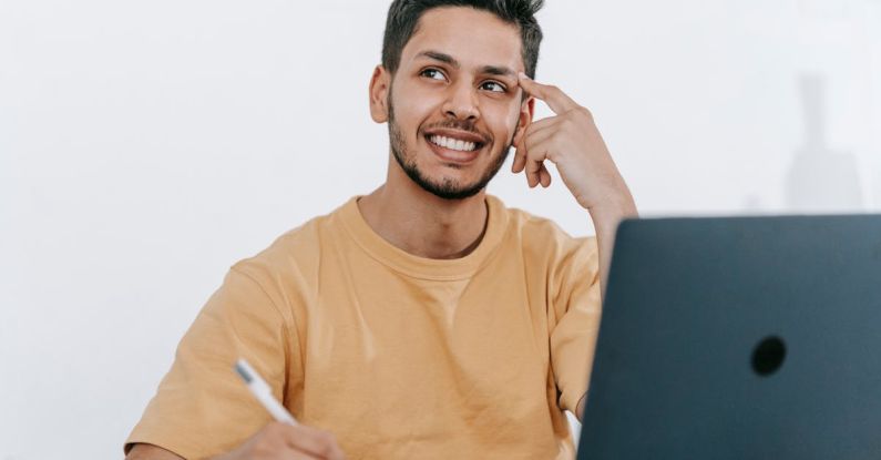 Work Future - Smiling young bearded Hispanic male entrepreneur thinking over new ideas for startup project and looking away dreamily while working at table with laptop and taking notes in notebook
