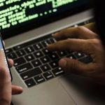 Malware Evolution - Unrecognizable hacker with smartphone typing on laptop at desk